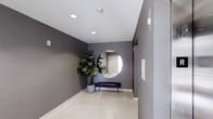 This single-floor space makes a strong first impression with its thoughtfully designed entryway. The corridor is finished with deep charcoal-toned walls that create a bold, modern aesthetic, complemented by light-colored flooring that brightens the area and adds a sense of openness. Recessed ceiling lighting provides clean, even illumination throughout, enhancing the contemporary feel of the space.

A standout design feature is the large, round wall mirror that serves as a striking focal point, reflecting light and adding depth to the entryway. Beneath it, a sleek, low-profile bench with metal legs offers both style and functionality. A lush fiddle-leaf fig tree in a modern planter adds a welcome touch of natural greenery, softening the space and introducing an organic element to the otherwise refined, minimal palette.

The right-hand wall features a textured finish, providing subtle visual contrast against the smooth charcoal surfaces, while an information panel mounted nearby adds a practical element to the design. The overall ambiance is polished and refined, balancing modern sophistication with welcoming touches — making it an ideal transitional space that sets the tone for what lies beyond.