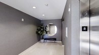 This single-floor space makes a strong first impression with its thoughtfully designed entryway. The corridor is finished with deep charcoal-toned walls that create a bold, modern aesthetic, complemented by light-colored flooring that brightens the area and adds a sense of openness. Recessed ceiling lighting provides clean, even illumination throughout, enhancing the contemporary feel of the space.

A standout design feature is the large, round wall mirror that serves as a striking focal point, reflecting light and adding depth to the entryway. Beneath it, a sleek, low-profile bench with metal legs offers both style and functionality. A lush fiddle-leaf fig tree in a modern planter adds a welcome touch of natural greenery, softening the space and introducing an organic element to the otherwise refined, minimal palette.

The right-hand wall features a textured finish, providing subtle visual contrast against the smooth charcoal surfaces, while an information panel mounted nearby adds a practical element to the design. The overall ambiance is polished and refined, balancing modern sophistication with welcoming touches — making it an ideal transitional space that sets the tone for what lies beyond.