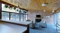 This inviting single-floor space combines rustic charm with comfortable, functional design. The standout architectural feature is the warm, tongue-and-groove wood-paneled ceiling, which lends a cozy cabin-like character to the interior. Ceiling fans are suspended throughout, ensuring comfortable airflow across the space.

A full wall of large windows floods the room with abundant natural light while offering picturesque views of the tree-lined outdoor surroundings, including a fenced exterior area. The exposed brick accent wall serves as a dramatic focal point, framing a classic fireplace with a wood mantel, above which a flat-screen television is mounted — creating a natural gathering hub for relaxation and entertainment.

The lounge area is furnished with a tasteful arrangement of patterned armchairs and a sleek sofa, centered around a coffee table atop a bold blue geometric-patterned carpet that adds a modern contrast to the rustic elements. Lush hanging plants throughout the space bring a refreshing touch of greenery indoors, enhancing the welcoming ambiance.

A built-in counter or reception desk is visible at the foreground, suggesting a dual-purpose layout suited for community gatherings, events, or hospitality. Overall, the space masterfully balances natural materials, ample light, and comfortable furnishings.
