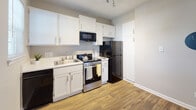 Located at 703 Grathwol Dr in Wilmington, NC, this single-floor unit offers a practical and well-maintained layout with a cohesive design throughout. The living area is characterized by rich, dark hardwood-style flooring, neutral warm-toned walls, and white baseboards that create a clean, polished look. A ceiling fan overhead and a window that brings in natural light contribute to a comfortable and airy atmosphere. The open flow between the living and adjacent spaces is enhanced by partial-height column dividers, adding a subtle architectural detail to the layout.

The kitchen is efficiently designed, featuring white beadboard-style cabinetry that extends to the upper walls for ample storage. It is equipped with stainless steel appliances, including a range and over-the-range microwave, along with a black refrigerator and dishwasher. Light wood-tone flooring and track lighting give the kitchen a bright, functional feel.

The bathroom presents a clean, white aesthetic with a wide vanity countertop, beadboard-style cabinetry beneath the sink, brushed nickel fixtures, and a well-lit mirror above. A shower with a curtain is visible in the reflection, completing the space. Overall, the unit provides a straightforward and move-in-ready environment suited for everyday living.
