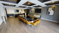 This single-floor space delivers a striking contemporary design centered around a bold contrast palette of black, white, and gold. The open-concept layout seamlessly connects a fully equipped kitchen to a spacious dining and lounge area, making it well-suited for gatherings and events.

The kitchen features dark cabinetry, a large island with bar seating, stainless steel appliances, and a range hood — offering both functionality and visual appeal. A white countertop island provides ample prep and serving space.

The dining area is anchored by a sleek, large-format black table surrounded by plush amber/gold upholstered chairs with gold-toned legs, creating a warm yet sophisticated focal point. The dark accent wall hosts a wall-mounted flat-screen television alongside a floating white credenza with decorative accessories.

Overhead, a dramatic coffered ceiling with black-trimmed recessed panels adds architectural depth and visual interest. Wood-look tile flooring runs throughout, grounding the space with warmth. Recessed lighting ensures the entire area is well-lit and inviting, contributing to the modern, upscale ambiance of the space.