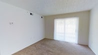 This single-floor residential space offers a clean, functional layout designed for comfortable everyday living. The main living area is finished with wall-to-wall carpeting in a neutral tan tone, providing a warm and soft underfoot feel that complements the light, neutral wall colors throughout. A textured popcorn ceiling adds a classic touch overhead, while recessed HVAC vents ensure consistent climate control throughout the space. A prominent sliding glass door fitted with vertical blinds serves as both a source of abundant natural light and a gateway to the outdoors, creating a bright and airy atmosphere in the living area. The vertical blinds offer flexible light control, allowing occupants to easily adjust the ambiance of the room. The walls feature standard electrical outlets positioned conveniently throughout, reflecting a practical and user-friendly design. The overall aesthetic is understated and versatile, providing a neutral canvas well-suited for a variety of furnishing styles and personal décor preferences. The single-floor layout promotes ease of movement and accessibility, making the space straightforward to navigate. With its emphasis on natural light, open feel, and simple finishes, this space presents a welcoming and livable environment.