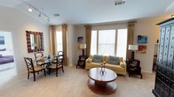 This single-floor space offers an inviting open-concept layout that integrates the living and dining areas into one cohesive environment. Large windows dressed with elegant floor-length golden drapes flood the interior with natural light, creating a warm and airy atmosphere throughout. The living area is anchored by a yellow sofa accented with blue throw pillows, complemented by a distinctive round wooden coffee table featuring a sculptural pedestal base. Track lighting along the ceiling provides additional illumination, while crown molding adds a refined architectural detail. The dining area accommodates a glass-top table surrounded by dark wood chairs with upholstered seats, ideal for intimate gatherings. Decorative lamps on side tables and a piece of wall art contribute to the polished, coordinated aesthetic. Neutral-toned carpeting runs throughout, softening the space and tying the warm color palette together. Overall, the space presents a comfortable yet tasteful environment, balancing functionality with classic design elements.