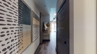 This single-floor unit showcases an industrial-modern aesthetic defined by exposed concrete ceilings and visible steel ductwork that run through the living area, lending the space a distinctly urban loft character. Hardwood-style flooring flows throughout, tying the rooms together cohesively.

The entry corridor sets an artistic tone, featuring curated wall art and a freestanding mirror with a leaning storage ladder, leading naturally into the main living space. Floor-to-ceiling windows in the living room flood the area with natural light and provide views of the surrounding exterior, while sheer drapery softens the industrial elements. The living room comfortably accommodates a seating arrangement alongside a media console area.

The kitchen features white shaker cabinetry with matte black hardware, granite-style countertops, a subway tile backsplash, and a full suite of stainless steel appliances including a range, refrigerator, microwave, and dishwasher.

The bedroom features a muted green accent wall, a concrete ceiling detail, and a space-saving barn door that reveals a stacked washer and dryer unit tucked neatly within the room. The bathroom offers a tub-shower combination with large-format tile surround and a granite-topped vanity.