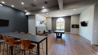 This single-floor recreational lounge is designed as a multifunctional social space, blending game amenities with comfortable gathering areas in a cohesive, modern aesthetic. The room is anchored by warm-toned hardwood flooring that runs throughout, complementing a color palette of dark charcoal, soft grey, and crisp white walls accented by gold and striped decorative elements.

The gaming area is well-equipped, featuring a full-size pool table as a central focal point, a shuffleboard table positioned along one wall, and a dart board mounted nearby. Wall-mounted flat-screen televisions provide entertainment options throughout the space. A prominent arched window along one wall allows natural light to filter in while offering views of the surrounding greenery outdoors.

Seating is varied and thoughtfully arranged. High-top bar tables with tan leather-style chairs provide casual dining or social seating, while a tufted booth-style banquette creates a more intimate lounge area. Small café-style tables and chairs are arranged near the arched window, forming a relaxed conversation zone.

A built-in wet bar or service counter with dark navy cabinetry and recessed accent lighting adds functional elegance. Decorative wood slab wall art, framed aviation-themed prints, and bold geometric wall accents contribute to the space's distinctive personality. Recessed ceiling lighting throughout ensures well-distributed illumination across all zones.