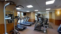 Located at 1910 Hawks Ridge Dr in Verona, Wisconsin, this fitness center offers a well-equipped, single-floor exercise facility designed to support a comprehensive workout experience. The space features a drop-panel ceiling with recessed fluorescent lighting that ensures consistent, bright illumination throughout. Large windows along the walls allow natural light to fill the room, creating an open and energizing atmosphere.

The cardio section is prominently equipped with multiple treadmills and an elliptical trainer, each positioned to take advantage of the natural light and window views. A wall-mounted television is conveniently placed for entertainment during workouts. Full-length mirrors line one wall, visually expanding the space and allowing users to monitor their form during exercise.

The strength training area features a multi-station cable and weight machine, along with free weight equipment, providing options for a full-body resistance workout. Rubber floor mats are placed beneath the equipment to protect the flooring and provide stability during use. The warm, neutral-toned wall paneling and wainscoting add a polished, welcoming aesthetic to the functional space. Overall, the facility is thoughtfully laid out to accommodate both cardio and strength training needs in a clean, comfortable environment.