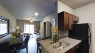This single-story residence is designed with an open-concept floor plan that seamlessly connects the kitchen, dining, and living areas. The kitchen serves as a central hub, showcasing rich dark-wood cabinetry with brushed nickel hardware, granite countertops, a stainless steel undermount sink, and a breakfast bar with seating. Dark hardwood flooring extends throughout the main living areas, adding warmth and continuity to the space.

The dining area sits adjacent to the kitchen, accommodating a full dining set within the open layout. The living room is generously proportioned, featuring a ceiling fan, recessed lighting, and a sliding glass door that opens to a private balcony, allowing natural light to fill the room.

The bedroom offers plush carpeting, a ceiling fan, and a closet with built-in shelving and hanging rods. A dedicated workspace area adds functional versatility to the room. The bathroom features a dual-sink vanity with matching granite countertops, dark cabinetry, a large mirror, and stylish wall-mounted lighting. Neutral gray walls throughout the residence create a cohesive, modern aesthetic that complements every room.