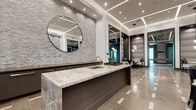 This upscale amenity space is defined by its contemporary design, premium finishes, and thoughtfully curated zones for relaxation and recreation. The central lounge area commands attention with soaring floor-to-ceiling windows that flood the space with natural light and offer sweeping views of the outdoor surroundings. A dramatic marble fireplace wall serves as a striking focal point, complemented by plush seating arrangements and refined soft furnishings.

The bar and kitchen area features a generously sized stone-topped island with a built-in sink, dark cabinetry, and a textured stacked-stone accent wall adorned with a large circular mirror. Recessed lighting and layered ceiling details enhance the polished atmosphere throughout. Dual flat-screen televisions are mounted above additional cabinetry, creating an entertainment-ready setting.

A dedicated billiards room, accessed through a dramatic dark-framed entryway, offers an intimate game space with accent lighting and bold, moody finishes. Flanking the entrance are upholstered booth-style seating areas, ideal for casual conversation. The fitness center, accessible through glass-paneled doors, is well-equipped and bright. Underfoot, large-format tile flooring with inlaid accent patterns ties the entire space together with a sense of architectural continuity.