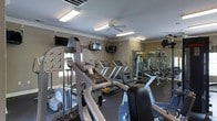 Located at 1025 Kensington Dr in Jacksonville, NC, this fitness center is a well-appointed, single-floor facility designed to support a comprehensive workout experience. The space is thoughtfully laid out to accommodate both cardio and strength training needs. A row of treadmills and step machines lines the floor, providing ample cardio options for users of all fitness levels. Complementing these are multi-station strength training machines, offering a full range of resistance and weight-based exercises. A large wall mirror spans a portion of the room, visually expanding the space and allowing users to monitor their form during workouts. The facility is well-lit with bright overhead fluorescent lighting and a ceiling fan that helps maintain a comfortable environment during intense sessions. Multiple mounted flat-screen televisions are positioned throughout the room, ensuring entertainment is always within view. Crown molding along the upper walls adds a subtle architectural touch, lending the space a polished appearance. The neutral-toned walls and dark flooring create a clean, focused atmosphere conducive to exercise. Overall, this fitness center is a practical and fully equipped space suitable for a wide range of fitness routines.