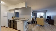 This thoughtfully designed single-floor residence offers a comfortable and functional layout across several well-defined living spaces. The open-concept living and dining area forms the heart of the home, featuring light wood-style flooring, neutral walls, and natural light streaming in through large windows. The living room accommodates a full seating arrangement and is complemented by an adjacent dining area with room for a four-person table and contemporary wall art.

The kitchen is efficiently laid out with dark cabinetry, granite-style countertops, white appliances, and a built-in pantry with open shelving for additional storage. A peninsula with a breakfast bar and an under-counter dishwasher connects the kitchen to the main living space, offering both practicality and casual dining options.

The primary bedroom features plush carpeting, a large window with natural light, and space for a full bedroom suite. A secondary room, carpeted and windowed, offers flexibility as a bedroom or home office. The bathroom is a standout feature, boasting a generous double-sink vanity with dark cabinetry, granite-style countertops, a wide mirror, and wood-style flooring. Throughout, the space maintains a cohesive, contemporary aesthetic with a calm, neutral color palette.