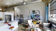 This single-level Dallas loft showcases an striking industrial aesthetic defined by soaring ceilings that amplify the sense of openness throughout the space. Exposed ductwork runs prominently overhead, complemented by a ceiling fan and pendant lighting suspended on long cords — hallmark features of converted industrial architecture. Structural concrete columns add to the raw, urban character of the interior.

The living area features polished concrete floors that reflect natural light and serve as a durable, visually cohesive foundation. A carefully arranged seating area includes a sculptural white tufted accent chair paired with a glass-top coffee table and a layered area rug, creating a sophisticated focal point. The walls are kept in a clean, neutral grey palette, providing an ideal backdrop for eclectic artwork, including a bold automotive photograph and a convex mirror that add depth and personality.

A console table styled with decorative objects, a bar cart, and various curated accents contribute to the loft's gallery-like ambiance. The kitchen area, partially visible, features clean white surfaces that contrast with the industrial surroundings. Overall, the space masterfully balances raw architectural elements with refined, contemporary furnishings.