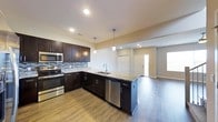 This contemporary 2-floor residence is thoughtfully designed to blend style and functionality throughout. The kitchen serves as a standout centerpiece, showcasing rich dark espresso cabinetry that extends to the ceiling, complemented by striking granite countertops with subtle veining. A peninsula with an undermount sink adds both prep space and a natural gathering point, while pendant lighting above provides warm, focused illumination. The backsplash features a mosaic tile pattern in cool blue and grey tones, adding a layer of visual texture to the space. Stainless steel appliances, including a range, microwave, and dishwasher, complete the modern kitchen aesthetic. Recessed lighting throughout ensures the space remains bright and well-lit. The kitchen flows seamlessly into an open living area, connected by warm hardwood-style flooring that runs throughout the main level. The living space benefits from natural light streaming in through windows near the entryway, enhancing the home's airy and welcoming ambiance. Clean white walls and neutral tones create a versatile backdrop, while the open layout between rooms promotes a sense of spaciousness. Spanning two floors, this residence offers a well-organized and modern living environment suitable for a variety of lifestyles.