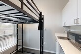 Located at 6105 Roosevelt Way NE in Seattle's Roosevelt neighborhood, this efficiently designed studio unit makes the most of its compact footprint through thoughtful layout and modern finishes. The entrance opens into a narrow hallway with a dedicated closet featuring a wire shelving system, providing practical storage from the moment you step inside. Dark hardwood-style flooring flows throughout, grounding the space with a contemporary warmth. The main living area is a well-considered combination of sleeping, living, dining, and kitchen functions. A loft-style bed frame with a built-in ladder maximizes vertical space, freeing up the floor below for a workspace positioned beneath a horizontal window that filters in natural light. The kitchen includes white upper cabinetry, a light countertop, and a stainless steel sink, offering clean, functional utility. Neutral gray walls throughout maintain a calm, modern aesthetic. The bathroom is finished with dark wood-toned cabinetry, a rectangular vessel-style sink with a modern faucet, a large mirror, and a separate shower and toilet area with built-in grab bars and tile-effect flooring. The overall design is clean, minimal, and purposefully organized to support comfortable everyday living in a compact urban setting.