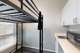 Located at 6105 Roosevelt Way NE in Seattle's Roosevelt neighborhood, this efficiently designed studio unit makes the most of its compact footprint through thoughtful layout and modern finishes. The entrance opens into a narrow hallway with a dedicated closet featuring a wire shelving system, providing practical storage from the moment you step inside. Dark hardwood-style flooring flows throughout, grounding the space with a contemporary warmth. The main living area is a well-considered combination of sleeping, living, dining, and kitchen functions. A loft-style bed frame with a built-in ladder maximizes vertical space, freeing up the floor below for a workspace positioned beneath a horizontal window that filters in natural light. The kitchen includes white upper cabinetry, a light countertop, and a stainless steel sink, offering clean, functional utility. Neutral gray walls throughout maintain a calm, modern aesthetic. The bathroom is finished with dark wood-toned cabinetry, a rectangular vessel-style sink with a modern faucet, a large mirror, and a separate shower and toilet area with built-in grab bars and tile-effect flooring. The overall design is clean, minimal, and purposefully organized to support comfortable everyday living in a compact urban setting.