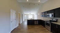 This single-floor space showcases a thoughtfully designed open-concept layout that effortlessly connects the kitchen and living areas. The kitchen is a standout feature, boasting dark, rich cabinetry paired with light stone countertops and a full suite of stainless steel appliances, including a range and dishwasher. A central island with a built-in sink serves as both a functional workspace and a natural gathering point. Recessed lighting and elegant pendant fixtures illuminate the kitchen, while a ceiling fan anchors the adjacent living area. Soaring vaulted ceilings amplify the sense of space throughout, creating an airy and expansive atmosphere. Rich hardwood flooring runs continuously across the open floor plan, adding warmth and cohesion to the interior. Natural light filters in through windows at the far end of the space, brightening the living area and enhancing the overall sense of openness. Additional doors along the hallway suggest well-appointed utility and storage spaces. The clean, neutral wall tones complement the bold cabinetry, resulting in a modern yet welcoming aesthetic throughout this well-appointed single-level home.