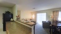 This single-floor residence is designed around an open-concept floor plan that promotes a seamless flow between its key living areas. A prominent breakfast bar with a stone-finished countertop serves as the central dividing feature between the kitchen and the combined dining and living space. The bar provides casual seating with dark bar-height chairs, making it a functional and social hub of the home.

Beyond the bar, the dining and living areas share a continuous carpeted space finished in neutral, warm tones. The living room is furnished with a light-toned sofa accented with decorative pillows, complemented by a patterned accent chair nearby. A floor lamp provides warm ambient lighting to the seating area.

A large sliding glass door dominates the far wall of the living room, fitted with vertical blinds that allow for adjustable natural light. The door provides both brightness and a connection to the outdoors. Walls throughout are painted in soft, neutral tones, and the ceilings are smooth with centrally placed ventilation. The overall ambiance is clean, understated, and comfortable.