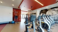The Retreat's fitness center is a thoughtfully designed, single-floor facility that combines functionality with a striking aesthetic. The space is immediately defined by its bold red ceiling accents, which contrast sharply with the clean white walls to create an energetic and motivating atmosphere. Linear LED lighting runs along the ceiling, providing bright, even illumination throughout the workout area.

The cardio section is outfitted with a lineup of Matrix brand equipment, including ellipticals, recumbent bikes, and upright cycles, arranged to allow comfortable spacing between machines. Beyond the cardio area, vertical wood slat dividers elegantly partition the space, leading into a dedicated strength and free weights zone. This area features a cable machine, a vertical knee raise station, a weight rack, and additional resistance training equipment, offering a comprehensive strength training experience.

Durable, rubber-textured flooring spans the entire facility, providing both safety and comfort during workouts. Natural light filters in through large windows at the far end of the gym, enhancing the open and airy feel of the space. The overall design balances bold visual elements with practical fitness amenities, making The Retreat's fitness center a well-rounded destination for residents seeking a complete workout experience.