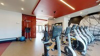 The Retreat's fitness center is a thoughtfully designed, single-floor facility that combines functionality with a striking aesthetic. The space is immediately defined by its bold red ceiling accents, which contrast sharply with the clean white walls to create an energetic and motivating atmosphere. Linear LED lighting runs along the ceiling, providing bright, even illumination throughout the workout area.

The cardio section is outfitted with a lineup of Matrix brand equipment, including ellipticals, recumbent bikes, and upright cycles, arranged to allow comfortable spacing between machines. Beyond the cardio area, vertical wood slat dividers elegantly partition the space, leading into a dedicated strength and free weights zone. This area features a cable machine, a vertical knee raise station, a weight rack, and additional resistance training equipment, offering a comprehensive strength training experience.

Durable, rubber-textured flooring spans the entire facility, providing both safety and comfort during workouts. Natural light filters in through large windows at the far end of the gym, enhancing the open and airy feel of the space. The overall design balances bold visual elements with practical fitness amenities, making The Retreat's fitness center a well-rounded destination for residents seeking a complete workout experience.