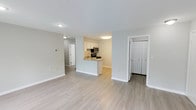 This single-floor residence offers a clean, modern aesthetic throughout, with light-toned luxury vinyl plank flooring unifying the main living areas. The open-concept layout connects the living room and kitchen seamlessly, with the living room bathed in natural light from a large window and sliding glass door fitted with sheer curtains. The kitchen is thoughtfully designed with white shaker-style cabinetry, granite-look countertops, a stainless steel appliance suite including a refrigerator, range, and microwave, and a peninsula that provides additional prep space and casual seating. The bedroom is generously sized, featuring plush wall-to-wall carpeting, a large closet with built-in wire shelving, and direct access to an en-suite bathroom complete with a single vanity, framed mirror, and a tub/shower combination with white tile surround. A second full bathroom offers similar finishes with a tub/shower and brushed nickel hardware. A dedicated laundry closet houses a full-size side-by-side washer and dryer. The space is finished with crisp white trim, neutral gray walls, and consistent ceiling-mount lighting throughout, creating a bright and cohesive living environment.