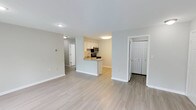 This single-floor residence offers a clean, modern aesthetic throughout, with light-toned luxury vinyl plank flooring unifying the main living areas. The open-concept layout connects the living room and kitchen seamlessly, with the living room bathed in natural light from a large window and sliding glass door fitted with sheer curtains. The kitchen is thoughtfully designed with white shaker-style cabinetry, granite-look countertops, a stainless steel appliance suite including a refrigerator, range, and microwave, and a peninsula that provides additional prep space and casual seating. The bedroom is generously sized, featuring plush wall-to-wall carpeting, a large closet with built-in wire shelving, and direct access to an en-suite bathroom complete with a single vanity, framed mirror, and a tub/shower combination with white tile surround. A second full bathroom offers similar finishes with a tub/shower and brushed nickel hardware. A dedicated laundry closet houses a full-size side-by-side washer and dryer. The space is finished with crisp white trim, neutral gray walls, and consistent ceiling-mount lighting throughout, creating a bright and cohesive living environment.