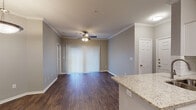 This one-bedroom, one-bathroom unit at The Humber in Little Rock, Arkansas offers a thoughtfully designed, single-story open-concept layout. The living area is finished with rich, dark wood-style flooring that extends throughout the main living spaces, complemented by crown molding and a ceiling fan overhead. A large sliding glass door dressed with vertical blinds allows natural light to fill the room and provides access to the outdoors.

The kitchen is a standout feature, centered around a peninsula with a granite countertop and an undermount double sink. White cabinetry provides ample storage, while stainless steel appliances—including a refrigerator, electric range, and microwave—add a modern touch. Subway tile backsplash and pendant lighting complete the space. A dedicated nook adjacent to the kitchen offers additional cabinetry and counter space.

The bedroom is carpeted for comfort, with a ceiling fan and double doors leading to a hallway that connects to additional storage. The bathroom features a granite-topped white vanity, wood-style flooring, brushed nickel fixtures, and a tiled tub-shower combination. Throughout the unit, neutral gray walls create a cohesive and versatile aesthetic.