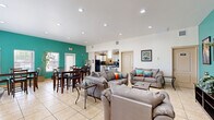 This single-floor community space offers a well-organized and functional layout designed to accommodate both relaxation and social gatherings. The main area is anchored by a spacious open-plan lounge and dining zone, unified by light beige ceramic tile flooring that runs throughout the entire space. A striking teal accent wall serves as the defining architectural feature, adding vibrant color contrast against the crisp white walls and ceilings. Vaulted ceilings in portions of the space enhance the sense of openness, while recessed lighting ensures the interior remains bright and inviting.

The lounge area is furnished with multiple plush gray sofas and loveseats accented with colorful throw pillows, arranged around a central coffee table and a wall-mounted flat-screen television set against the teal feature wall. A dedicated dining zone accommodates several high-top tables and bar-height chairs, bathed in natural light from multiple windows overlooking the exterior.

The kitchen is fully equipped with white cabinetry, dark granite countertops, black appliances including a refrigerator and range, and track lighting overhead. A connected entryway area features a frosted glass door, a decorative mirror, and a multi-function printer station. Tall potted trees and greenery are placed throughout, adding a natural, welcoming ambiance to the overall space.