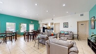 This single-floor community space offers a well-organized and functional layout designed to accommodate both relaxation and social gatherings. The main area is anchored by a spacious open-plan lounge and dining zone, unified by light beige ceramic tile flooring that runs throughout the entire space. A striking teal accent wall serves as the defining architectural feature, adding vibrant color contrast against the crisp white walls and ceilings. Vaulted ceilings in portions of the space enhance the sense of openness, while recessed lighting ensures the interior remains bright and inviting.

The lounge area is furnished with multiple plush gray sofas and loveseats accented with colorful throw pillows, arranged around a central coffee table and a wall-mounted flat-screen television set against the teal feature wall. A dedicated dining zone accommodates several high-top tables and bar-height chairs, bathed in natural light from multiple windows overlooking the exterior.

The kitchen is fully equipped with white cabinetry, dark granite countertops, black appliances including a refrigerator and range, and track lighting overhead. A connected entryway area features a frosted glass door, a decorative mirror, and a multi-function printer station. Tall potted trees and greenery are placed throughout, adding a natural, welcoming ambiance to the overall space.