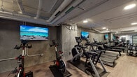 This fitness center offers a comprehensive workout environment designed to accommodate a wide range of exercise routines. The space features an industrial aesthetic, characterized by exposed piping along the ceilings, concrete-textured walls, and wood-look flooring throughout, lending the gym a modern yet raw character.

The cardio zone is well-stocked with Life Fitness treadmills, elliptical machines, a rowing machine, and stationary spin bikes, each positioned on protective rubber mats. Multiple wall-mounted flat-screen televisions are strategically placed throughout the cardio area, providing entertainment during workouts.

A dedicated strength training section houses a rack of dumbbells in varying weights, a weight bench, and a cable pulley machine, offering users the tools necessary for a full resistance training session. Rubber flooring tiles in this area provide safety and stability. A mini-split air conditioning unit ensures climate control in the strength zone.

An additional area is dedicated to functional fitness, featuring stability balls, medicine balls, kettlebells, resistance bands, and yoga mats, making the space versatile for stretching, core work, and dynamic training. Recessed lighting throughout ensures the entire facility is well-lit. The gym presents a practical, fully outfitted fitness environment suitable for a broad range of users and training styles.