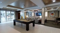 This single-floor amenity lounge is a well-appointed social and recreational space designed with modern elegance. At its center stands a sleek, dark-wood billiards table set atop light hardwood-style flooring, immediately drawing attention as the focal point of the room. The space is divided into distinct functional zones that flow seamlessly into one another. A generous lounge area features plush, neutral-toned sofas arranged around a wall-mounted flat-screen television, creating an inviting setting for relaxed gatherings. Accent lighting from stylish pendant and chandelier fixtures adds warmth and sophistication throughout, complementing the tray ceilings that lend an architectural depth to the interior. Structural columns are incorporated smoothly into the design, separating areas without interrupting the open, airy feel of the space. A dining area with dark wood furniture is visible toward the rear, offering additional seating options. Natural light filters in through large windows along one side, providing views of the exterior greenery and brightening the overall atmosphere. The neutral color palette, accented with warm tones and carefully curated artwork on the walls, gives the space a refined yet welcoming ambiance suited for both casual recreation and social events.