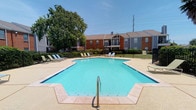 This single-story community amenity space offers two key facilities designed for recreation and wellness. The outdoor pool area serves as a central gathering point, featuring a generously sized, geometrically shaped pool with clear blue water and a wide concrete sundeck surrounding it. Multiple lounge chairs are arranged along the perimeter, providing ample space for sunbathing and relaxation. The pool is enclosed by a black metal fence, with mature trees providing partial shade and a backdrop of brick and siding residential buildings visible beyond. The overall atmosphere is open and bright, benefiting from abundant natural sunlight.

The onsite fitness center is housed in a well-lit, single-floor room with light-colored walls and textured ceilings. Large floor-to-ceiling mirrors line one wall, visually expanding the space and allowing users to monitor their form during workouts. The facility is equipped with a multi-station cable weight machine, a treadmill, an elliptical, and additional cardio equipment. Durable, low-pile flooring covers the exercise area, and French-style glass panel doors provide natural light and direct access to the outdoor grounds. A fire extinguisher and sanitizing station are also present, reflecting attention to safety and cleanliness.