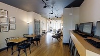 This single-floor space is thoughtfully arranged to accommodate multiple functional zones within an open, flowing layout. The interior features warm hardwood flooring that runs throughout, lending a cohesive and inviting character to the entire space. Track lighting mounted on a textured dark ceiling provides flexible, directed illumination across the room, complementing additional floor lamps that contribute a softer ambient glow.

Near the entrance, a casual dining area is furnished with wooden tables and black metal chairs, ideal for individual or small-group use. A paneled gray door adds a subtle architectural detail to this section of the room. Moving deeper into the space, the layout opens into a more relaxed lounge area, where low-profile seating and accent chairs create a comfortable, informal atmosphere. Natural light filters in through windows at the far end, brightening the back of the space and enhancing its open feel.

A partial partition wall on the right side of the room defines a workstation or service counter area equipped with monitors, suggesting a reception or operational function. Overall, the space balances a professional yet relaxed ambiance, suitable for a café, coworking environment, or community gathering space.