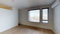 This single-floor space showcases a clean, contemporary design with a neutral color palette that creates a bright and airy atmosphere. The room is finished with light hardwood flooring that runs throughout, adding warmth to the otherwise minimalist aesthetic. Crisp white walls and a smooth white ceiling enhance the sense of openness and allow natural light to take center stage.

Two generously sized windows, each fitted with adjustable horizontal blinds featuring sleek black frames, dominate one wall and flood the space with abundant natural light. These windows offer elevated views of the surrounding urban environment, including notable city buildings and streetscapes, providing a dynamic urban backdrop.

A built-in open shelving unit is integrated into one corner of the room, offering practical storage and display space without compromising the clean lines of the interior. The shelves span floor to near-ceiling height, maximizing vertical space efficiently.

The overall ambiance is modern and understated, well-suited for a variety of uses. The combination of natural light, urban views, hardwood flooring, and thoughtful built-in storage makes this a versatile and functional space with strong contemporary appeal.