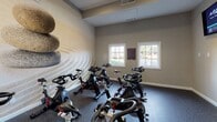 Located at 980 Walther Blvd in Lawrenceville, Georgia, this single-floor spin studio offers a focused and well-appointed environment designed for indoor cycling workouts. The room is furnished with a row of Matrix stationary spin bikes arranged to face a striking, large-scale Zen-inspired mural depicting stacked stones against a raked sand background, creating a calming focal point that balances the energy of the workout space. The flooring consists of durable, speckled rubberized material well-suited for high-activity use. Natural light enters through multiple white-framed windows along one wall, providing views of the surrounding outdoor greenery and brightening the interior. A wall-mounted mini-split HVAC unit ensures climate comfort throughout sessions. Two wall-mounted flat-screen televisions are positioned for easy viewing from the bikes, keeping users engaged and informed during rides. A sleek, freestanding digital kiosk station is also present, likely offering workout tracking or scheduling functionality. The studio connects to an adjacent fitness area through glass-paneled doors, suggesting it is part of a broader amenity facility. Neutral gray wall tones and clean white trim complete the polished, modern aesthetic of this dedicated cycling space.