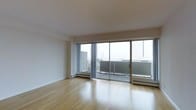 This well-appointed single-floor space showcases a clean, contemporary design throughout. The main living area is immediately striking, characterized by smooth white ceilings, crisp white walls, and warm light hardwood flooring that runs seamlessly across the room. Natural light pours in abundantly through a wall of expansive floor-to-ceiling sliding glass doors, which open onto a private balcony offering impressive views of the surrounding urban skyline. The large-panel glass doors create a strong visual connection between the interior living space and the outdoor environment, enhancing the sense of openness and airiness throughout. Baseboard heating units are integrated neatly beneath the windows, ensuring comfort while maintaining the space's streamlined aesthetic. The overall ambiance is minimalist and airy, with a neutral color palette that provides a versatile canvas for a variety of interior styles. The balcony adds valuable outdoor living space, framed by a modern glass railing that preserves the panoramic city views. This space is well-suited for those who appreciate modern design, abundant natural light, and elevated urban living.