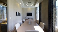 This elegant conference room is designed to accommodate professional meetings in a refined, modern setting. The space is anchored by a large rectangular conference table surrounded by sleek white chairs, providing seating for a full team. A tray ceiling adds architectural depth overhead, while a striking circular ring pendant light serves as a central focal point, casting a warm and even glow throughout the room. Recessed lighting further enhances the bright, polished atmosphere.

The walls are finished in a soft, textured neutral tone that conveys a calm and focused ambiance, complemented by subtle framed artwork that adds a touch of understated sophistication. A wall-mounted flat-screen display is positioned at the head of the room on a built-in credenza, making presentations and collaborative work seamless. Integrated cable management on the conference table supports connectivity for multiple users.

Natural light fills one side of the room through tall windows dressed with patterned drapery, adding warmth and visual interest while maintaining privacy as needed. The overall design strikes a balance between functionality and elegance, making this conference space well-suited for executive meetings, client presentations, or collaborative work sessions.