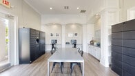 Located at Grandewood Pointe in Orlando, Florida, this community amenity space serves as both a business center and a package/mail room. The single-floor layout is thoughtfully designed with a clean, contemporary aesthetic, highlighted by crisp white walls, elegant crown molding, and arched architectural niches that add a refined, classic character to the interior.

The main area centers around a large granite-topped communal table with industrial-style metal chairs, providing a comfortable space for collaborative work or casual use. Flanking the table are built-in workstations tucked within the arched alcoves, each equipped with a desktop computer, offering residents convenient access to productivity tools. A printer station and additional cabinetry with granite countertops add to the functional layout.

Two Luxer One smart package locker systems are prominently featured — one a full-height tower unit and another a multi-bay wall-mounted system — enabling secure, 24/7 parcel retrieval. Light wood-look flooring runs throughout, complementing the neutral warm-beige and white color palette.

Large multi-pane windows along the perimeter flood the space with abundant natural light while framing views of the surrounding landscaped grounds. Recessed lighting supplements the natural illumination, ensuring a bright and welcoming atmosphere throughout the day.
