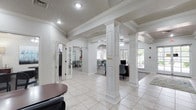 This well-appointed single-floor space makes a strong first impression with its grand, open layout anchored by a series of decorative white columns and graceful arched passageways that define each area while maintaining a seamless, connected flow. The main reception area features a rich dark wood desk accompanied by houndstooth-patterned accent chairs, setting a polished yet inviting tone. Light tile flooring runs through the main corridors and gathering areas, complemented by tasteful area rugs that add warmth and texture underfoot. Coffered and tray ceilings with crown molding add architectural depth, while recessed lighting and ceiling fans ensure the space remains bright and comfortable. Elegant arched openings lead to a dedicated dining or meeting area furnished with round tables and upholstered seating in soft teal tones, alongside colorful wall art that lends a vibrant, curated aesthetic. Large windows at the far end of the space invite generous natural light and offer views of the surrounding greenery. Additional rooms accessible through French doors provide flexible spaces for private meetings or workstations. The overall ambiance is clean, classic, and professional, combining traditional architectural character with a light, airy atmosphere suited for community gatherings or professional engagements.