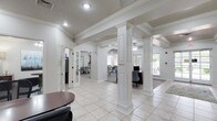 This well-appointed single-floor space makes a strong first impression with its grand, open layout anchored by a series of decorative white columns and graceful arched passageways that define each area while maintaining a seamless, connected flow. The main reception area features a rich dark wood desk accompanied by houndstooth-patterned accent chairs, setting a polished yet inviting tone. Light tile flooring runs through the main corridors and gathering areas, complemented by tasteful area rugs that add warmth and texture underfoot. Coffered and tray ceilings with crown molding add architectural depth, while recessed lighting and ceiling fans ensure the space remains bright and comfortable. Elegant arched openings lead to a dedicated dining or meeting area furnished with round tables and upholstered seating in soft teal tones, alongside colorful wall art that lends a vibrant, curated aesthetic. Large windows at the far end of the space invite generous natural light and offer views of the surrounding greenery. Additional rooms accessible through French doors provide flexible spaces for private meetings or workstations. The overall ambiance is clean, classic, and professional, combining traditional architectural character with a light, airy atmosphere suited for community gatherings or professional engagements.