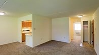 This single-floor space features an open-concept layout that connects the main living area to the kitchen, creating a cohesive and functional flow throughout. The living area is finished with wall-to-wall carpeting in a neutral tone, providing a comfortable underfoot feel and a clean, understated aesthetic. Smooth white walls and standard-height ceilings keep the space feeling bright and open.

The kitchen is partially separated by a half-wall partition that doubles as a breakfast bar or pass-through counter, maintaining visual connectivity between the two spaces. It is equipped with warm-toned wooden cabinetry, a white electric range, and a light-colored countertop, offering practical storage and workspace. Under-cabinet lighting adds a functional touch to the cooking area.

A hallway visible to the right of the living area leads to additional rooms, suggesting a well-organized floor plan with clear separation between living and private spaces. Natural light filters in from that direction, contributing to the overall brightness of the unit. The neutral color palette throughout the space provides a versatile backdrop suitable for a variety of interior styles.