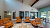 This single-story clubhouse is defined by its striking architectural design, highlighted by soaring vaulted ceilings with exposed dark wood beams and a white shiplap ceiling finish that create an open, airy atmosphere throughout. The main lounge area serves as the centerpiece of the space, featuring rich hardwood-style flooring, a cohesive arrangement of bold orange upholstered armchairs, and a patterned area rug anchoring the seating grouping around a low coffee table. Large floor-to-ceiling glass panels and sliding doors line the perimeter, flooding the interior with natural light and offering direct views of the outdoor pool and landscaped surroundings. An adjacent wing, painted in a calming teal-blue tone, provides additional flexible seating with bistro-height tables and stools positioned near oversized windows and doors that open directly to the exterior. The broader layout reveals additional functional areas, including what appears to be a private conference or meeting room with formal seating and a fitness area visible through interior glass partitions. Abstract artwork adorns the tall white walls, adding a contemporary touch. The overall ambiance is welcoming and social, balancing vibrant design elements with comfortable, community-oriented gathering spaces.