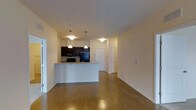 Located at 310 @ Nulu, this single-floor unit offers an open-concept layout that seamlessly connects the kitchen and living areas. The kitchen is outfitted with dark cabinetry, black appliances including a microwave and refrigerator, and a peninsula countertop with light granite-style surfaces that provides both workspace and a casual dining or bar seating option. Pendant lighting hangs above the peninsula, adding focused illumination and a stylish touch to the kitchen space.

The living area is generously proportioned, featuring warm hardwood-style flooring that runs throughout and contributes to the cohesive, modern aesthetic. Neutral cream-toned walls keep the space feeling open and bright, while flush ceiling lighting supplements the overall illumination. A hallway visible in the background leads to additional rooms, suggesting a practical and well-organized floor plan. The unit's clean lines, contemporary finishes, and efficient use of space make it a functional and attractive offering within the NuLu neighborhood.