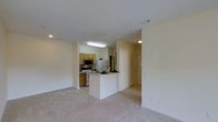 Located at 8800 Cattail Creek Pl in Raleigh, NC, this single-floor residence offers a well-organized, open layout designed for comfortable everyday living. The living area features neutral-toned wall-to-wall carpeting that creates a warm and inviting atmosphere, flowing seamlessly toward the kitchen and dining space.

The kitchen is equipped with warm wood-toned cabinetry, a full suite of appliances including a refrigerator, microwave, and range, as well as a decorative tile backsplash that adds visual interest. A peninsula-style breakfast bar extends from the kitchen, providing a casual dining or workspace option while maintaining an open connection to the living area.

The transition between the kitchen and living spaces is defined by a partial wall and passthrough, allowing natural sightlines throughout the unit. Overhead lighting in the kitchen ensures a well-lit workspace, while recessed lighting serves the living area. The hallway beyond the kitchen leads to additional rooms, with a closet featuring louvered doors visible in the background, adding practical storage to the layout. The overall ambiance is clean, neutral, and move-in ready, reflecting a functional and straightforward residential design.
