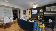 This single-floor residence offers a well-designed open-concept layout that seamlessly connects the living, dining, and kitchen areas. The living room serves as the centerpiece of the space, anchored by a bold dark accent wall that frames a wall-mounted flat-screen TV and a decorative arrangement of round mirrors in gold and blue tones. A plush blue sofa with accent pillows sits atop a patterned area rug, complemented by warm hardwood flooring that runs throughout. A ceiling fan with integrated lighting adds both function and a finishing touch to the room.

The kitchen is visible from the living area and features dark cabinetry, black appliances including a range and built-in microwave, and a breakfast bar with bar stools that provides a casual dining option. The kitchen's tile backsplash and functional layout make it well-suited for everyday cooking.

Adjacent to the main living space, a laundry area is conveniently accessible. The overall ambiance of the residence is modern and inviting, with a cohesive color palette of navy, gold, and charcoal that ties each area together. The open sightlines between rooms enhance the sense of space, making this a comfortable and visually engaging home.