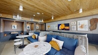 Located at 1501 Buena Vista Ave in Alameda, California, this single-floor amenity space seamlessly blends comfort, style, and functionality. The interior is defined by a striking warm wood plank ceiling that adds natural texture and warmth throughout, complemented by polished concrete floors and two-tone upholstered wall panels in soft gray and slate blue tones.

The lounge area features woven white rattan sofas dressed in rich blue cushions with mustard accents, arranged around marble-topped round tables — creating an inviting, resort-inspired social environment. A curated gallery wall with eclectic framed artwork and a large display screen anchors the space visually.

For focused work, a dedicated Wi-Fi lounge area offers a long built-in wooden countertop workstation with vibrant red and pink chairs set against an exposed brick accent wall, bathed in natural light from expansive floor-to-ceiling windows overlooking the street.

Private acoustic booths provide semi-enclosed seating with fully upholstered walls, built-in tables, and recessed lighting — ideal for calls or concentrated work sessions. Glass entry doors with aluminum frames connect the interior to the exterior, maintaining openness and accessibility. The overall ambiance is polished yet welcoming, balancing collaborative energy with quiet, productive retreats.