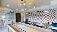 Located at 1701 Atria Ct in Raleigh, NC, this single-floor community space is thoughtfully designed with a contemporary aesthetic that balances comfort and visual interest. The lounge area immediately draws the eye with a bold geometric-patterned accent wall in deep burgundy and white, serving as a dramatic focal point that anchors the seating area. A cluster of Edison bulb pendant lights hang at varying lengths from the ceiling, casting a warm, inviting glow throughout the space and adding an industrial-chic character. A full-length shuffleboard table takes center stage on the floor, offering an engaging recreational feature for residents and guests. The seating arrangement includes round bistro-style tables paired with woven chairs, creating intimate gathering spots ideal for casual conversation or social events. Additional pendant globe lights complement the Edison fixtures, contributing to a layered lighting scheme that enhances the room's ambiance. The neutral ceiling and clean architectural lines provide a refined backdrop that allows the decorative elements to stand out. A dark-framed door and subtle teal accent wall visible in the background hint at thoughtful color coordination throughout. Overall, this space delivers a stylish, well-appointed lounge environment designed for community engagement and relaxation.