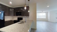 This single-floor space showcases a thoughtfully designed open-concept layout that connects the kitchen and living area in a clean, modern aesthetic. The kitchen is a standout feature, highlighted by a spacious granite-topped island with a double undermount stainless steel sink and a sleek pull-down faucet. Dark espresso cabinetry provides bold contrast against the light granite surfaces, while a classic white subway tile backsplash adds texture and visual interest. Black appliances, including a range and built-in microwave, complement the kitchen's contemporary palette. Two pendant lights with brass-toned fixtures hang above the island, providing focused task lighting with a stylish touch. The kitchen transitions naturally into the living area, which benefits from abundant natural light streaming through a large window fitted with horizontal blinds. Light-toned hardwood-style flooring runs throughout, contributing to a cohesive and open feel. A ceiling fan in the living area enhances comfort and airflow. The overall ambiance is clean, modern, and well-appointed, with a neutral color palette that creates a bright and welcoming environment throughout.