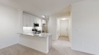 This single-story, 2-bedroom, 2-bathroom residence offers a thoughtfully designed layout with clean, contemporary finishes throughout. The open-concept living and kitchen area forms the heart of the space, where a peninsula with granite countertops serves as both a prep station and casual dining surface. The kitchen is well-equipped with a full suite of stainless steel appliances, including a range, microwave, refrigerator, and dishwasher, complemented by shaker-style cabinetry, under-cabinet lighting, and a tile backsplash. Wood-look flooring grounds the kitchen, while plush neutral carpeting extends through the living area and bedrooms. Both bedrooms are generously proportioned, each featuring a ceiling fan, carpeted floors, and natural light from windows with blinds. One bedroom includes vertical blinds suggesting access to a patio or larger window feature. The bathrooms are finished with wood-look tile flooring, granite-topped vanities, and tiled shower/tub combinations. A hallway connects the main entry to the private areas of the home, maintaining a clear and functional flow throughout this well-balanced, move-in ready residence.