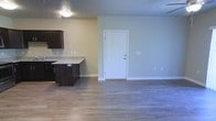 This single-floor space offers a clean, open-concept layout that integrates the kitchen and living area into one cohesive environment. The kitchen is appointed with dark espresso-toned cabinetry that provides a strong visual contrast against the light gray walls. A peninsula with a light-speckled granite-style countertop extends into the living space, offering additional prep space and a natural divider between the two functional zones. The flooring throughout is a warm-toned laminate wood that runs continuously across the entire space, contributing to a sense of openness and flow. The living area is generously proportioned, with ample room for a variety of furniture arrangements. A solid white entry door with paneled detailing serves as the primary access point, framed by clean white baseboards that run along the perimeter of the room. The walls are painted in a soft, neutral gray-green tone that creates a calm and welcoming ambiance. Flush-mount ceiling lighting provides consistent illumination throughout. The overall aesthetic is modern and understated, with a practical design that balances functionality and comfort in a well-maintained, move-in-ready condition.