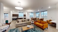 This single-floor communal space is thoughtfully designed to blend contemporary style with warm, inviting elements. The layout centers on an expansive open-plan lounge area, where wood-look tile flooring and crisp white walls create a clean, modern backdrop. A striking reclaimed wood feature wall runs the length of the space, adding rich texture and a rustic contrast to the otherwise contemporary aesthetic. A sleek, linear fireplace is set into this accent wall, anchoring the primary seating area, which is furnished with a neutral sofa, a cognac leather loveseat, and wood-topped coffee tables arranged atop a teal area rug. Large pendant lighting fixtures illuminate the space overhead. An abundance of natural light pours in through oversized windows along the perimeter, offering views of the surrounding landscape. The kitchenette area is well-appointed with dark wood cabinetry, white subway tile backsplash, light quartz countertops, a large-format island with bar seating, and a stainless steel sink. A dedicated dining nook, framed by corner windows, provides an intimate setting for small gatherings. The overall ambiance is welcoming and functional, combining residential comfort with a polished, communal character suited for shared use.