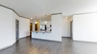 This single-floor space showcases a contemporary open-concept design that seamlessly integrates the kitchen and living areas into one cohesive layout. The kitchen is a standout feature, anchored by a peninsula with a granite-style countertop that doubles as a breakfast bar with seating. White upper cabinetry pairs cleanly with stainless steel appliances, including a French door refrigerator and built-in microwave, while pendant lights hang above the peninsula to provide focused task lighting and a modern aesthetic touch. The open layout allows the kitchen to flow naturally into the surrounding living space, creating a sense of connectivity and ease of movement throughout. Durable hardwood-style flooring in a warm gray tone runs consistently across the entire space, adding both visual continuity and a modern feel underfoot. The walls are finished in a crisp white, complemented by clean gray baseboards that frame each room with subtle contrast. Recessed and flush-mount ceiling lighting ensures the space remains well-illuminated. A hallway visible in the background suggests additional private rooms, while a sliding door to one side hints at further flexible use of space.